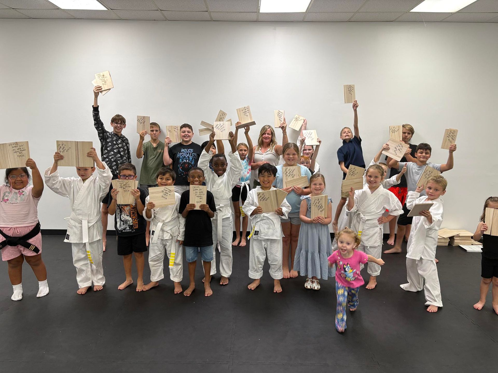 Group of children in martial arts attire holding up wooden boards in a studio.