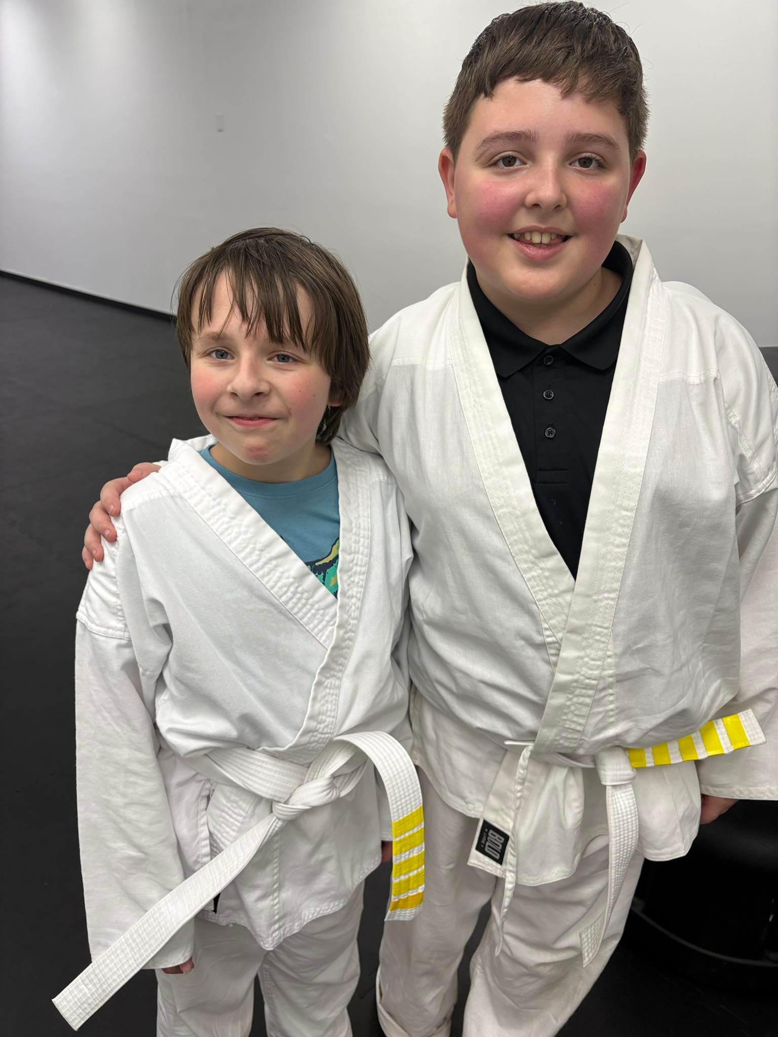 Two boys in white martial arts uniforms with yellow belts, posing indoors. One has arm around the other's shoulder.