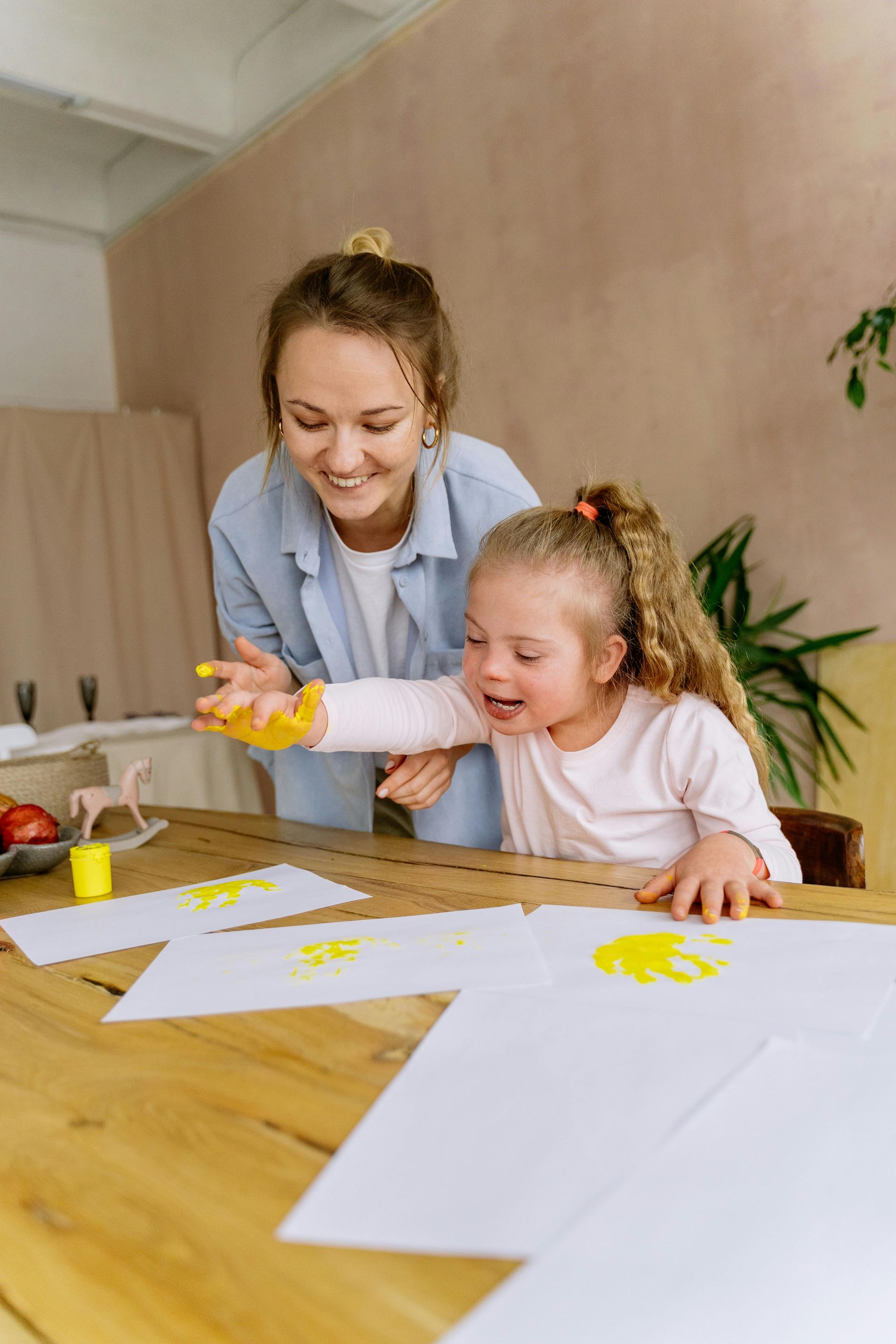 Woman assists a child with Down syndrome painting with yellow paint on paper at a table.