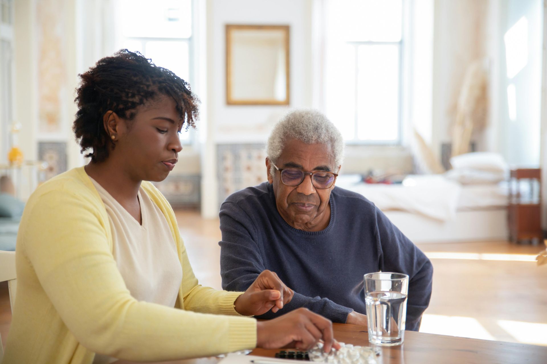 Woman assisting person with medication at a table; glass of water nearby.