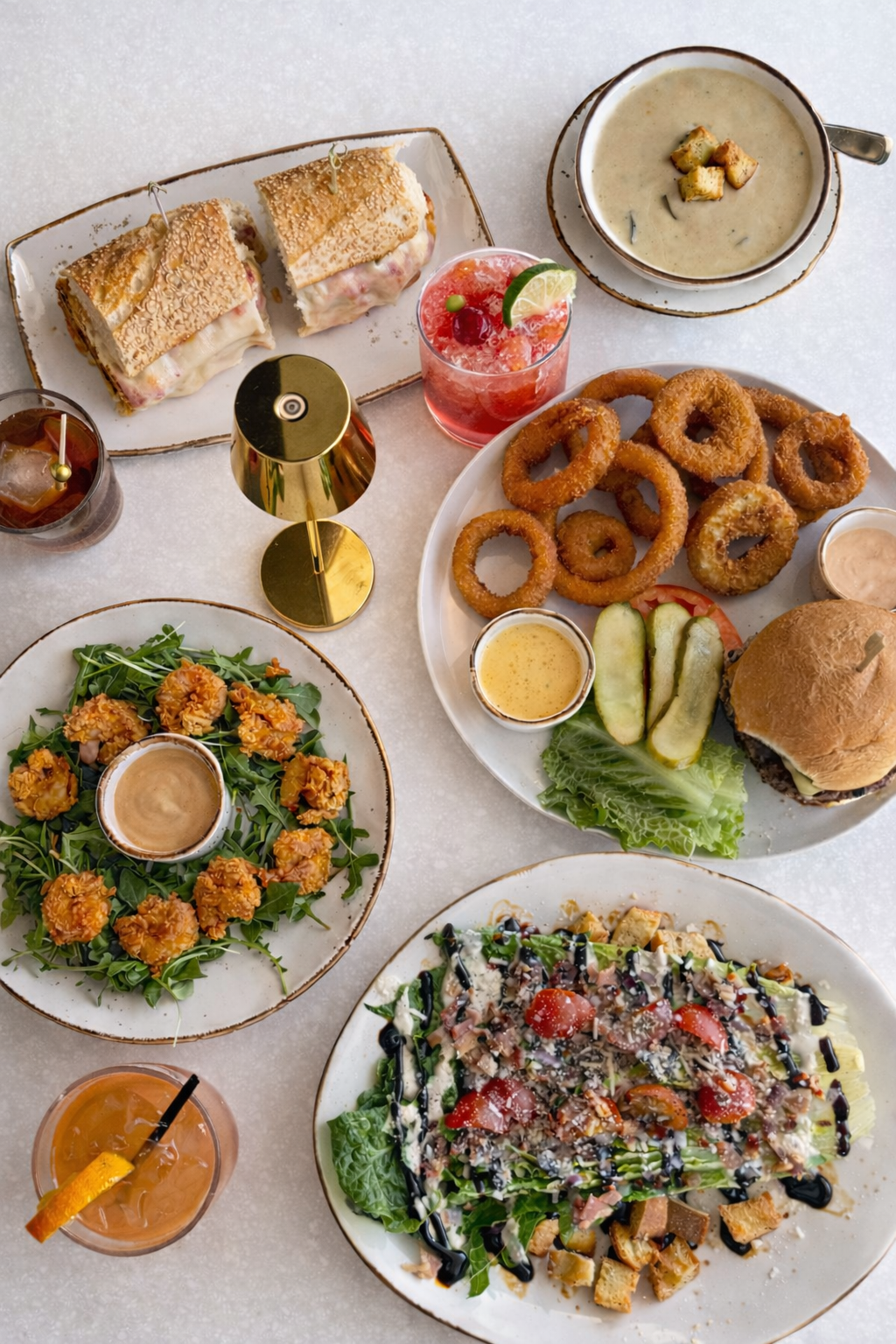 A flat-lay view of a table spread featuring a sandwich, soup, onion rings with a burger, salad, shrimp, and several drinks.