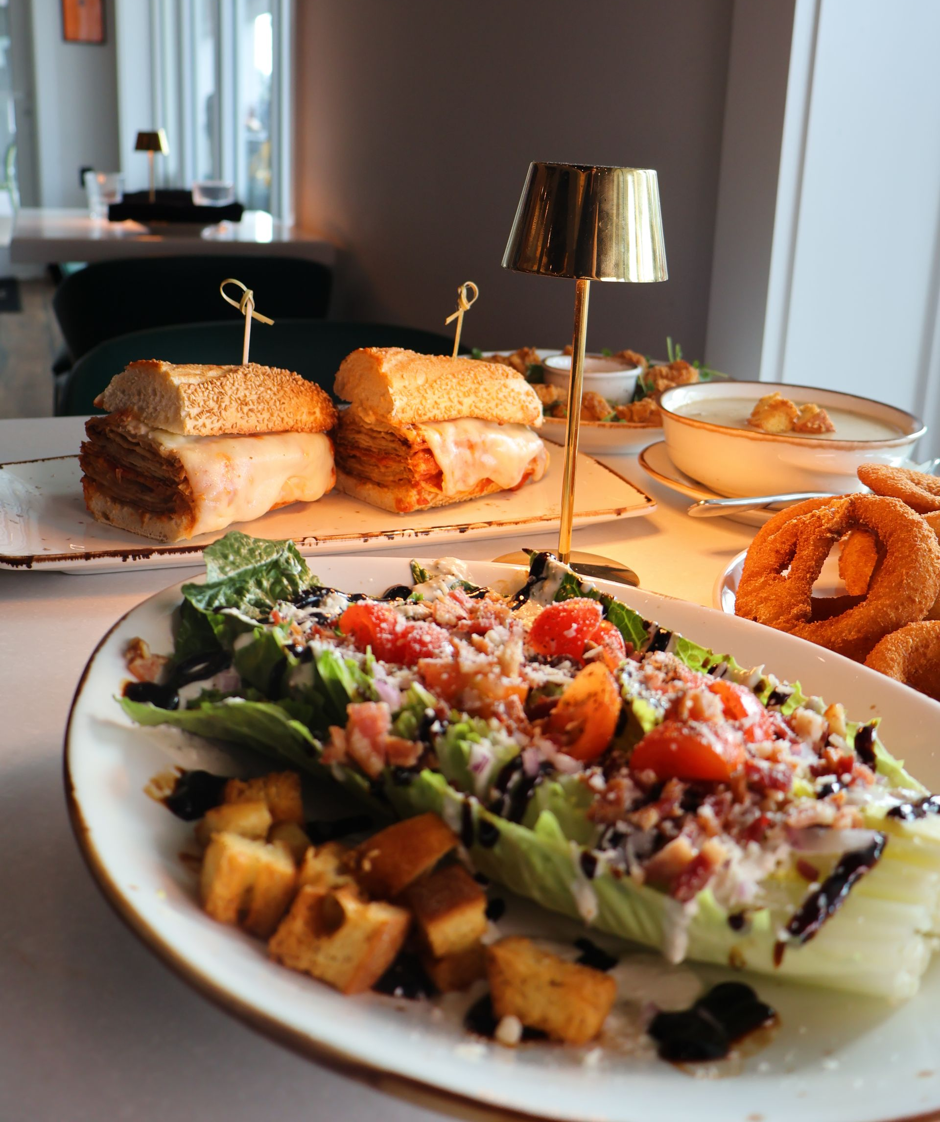 A table with a salad, sandwiches, onion rings, and soup, illuminated by a small gold lamp in a restaurant setting.