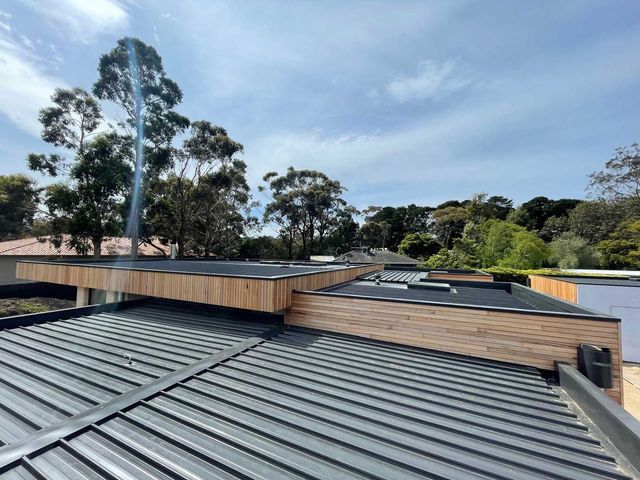 A view of a modern building roof featuring dark ribbed metal sections, wooden trim, and a smoking chimney against the sky.