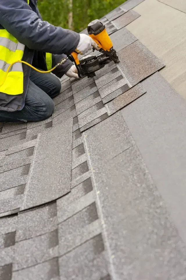 A roofer in a yellow safety vest using a pneumatic nail gun to install gray shingles on a sloped roof.