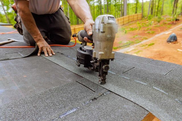 A worker uses a pneumatic nail gun to install asphalt shingles on a roof.
