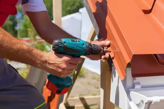 A person uses a teal cordless drill to fasten a piece of orange metal roofing near a white gutter on a construction site.