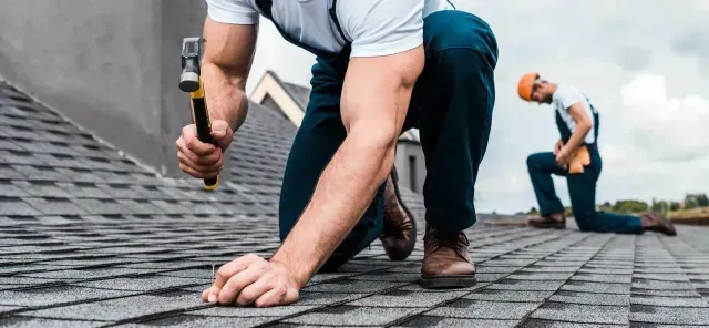 Two workers in workwear kneeling on a residential roof, installing gray asphalt shingles with a hammer.