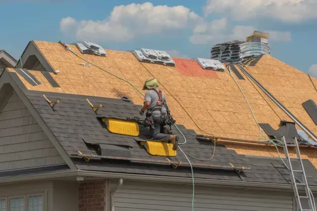 A construction worker wearing a safety harness installs shingles on a sloped residential roof under a bright blue sky.