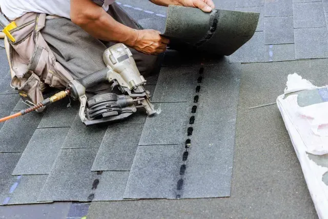 A construction worker uses a pneumatic nail gun to install grey asphalt shingles on a roof.