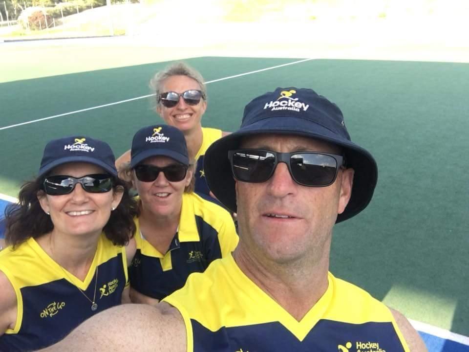 Group of four people wearing sunglasses and hats, smiling at the camera on a hockey field.