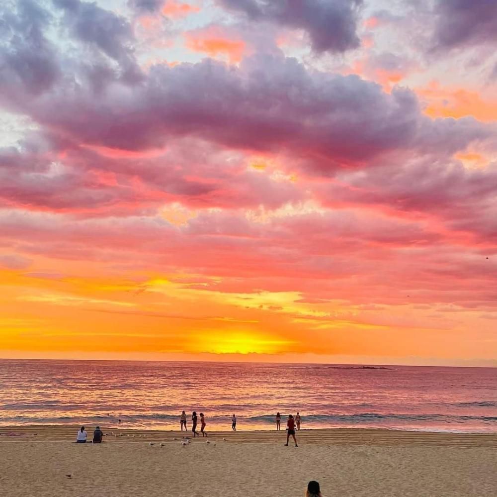 Sunset over the ocean, with vibrant orange, pink, and purple clouds. People on the beach.