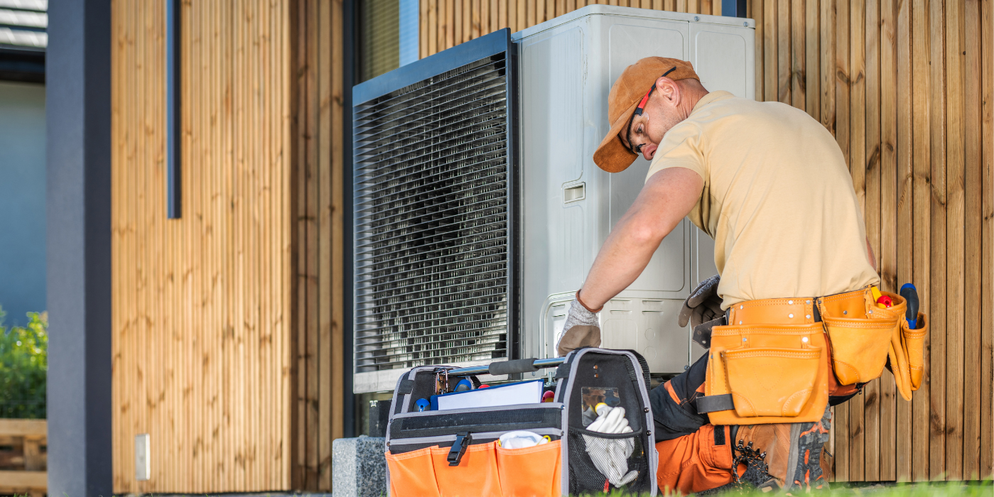 A repairman wearing a tool belt repairs an air conditioning unit outside a wooden building.