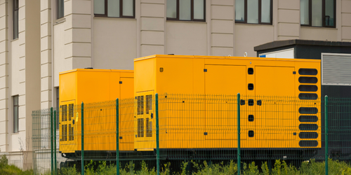 A row of yellow generators are parked in front of a building behind a fence.