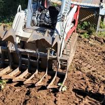 A white skid steer loader with a metal root rake attachment sits on churned, dark brown dirt in a bright outdoor setting.