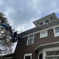 A worker in a blue boom lift performs maintenance on the exterior of a multi-story brown house with white trim.