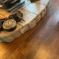 A stack of cast iron pans and a lidded Dutch oven sit on a curved, textured stone fireplace hearth above wood flooring.