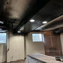 A room with smoke-damaged black ceilings, recessed lighting, white walls, and a wooden wall featuring a large cross.