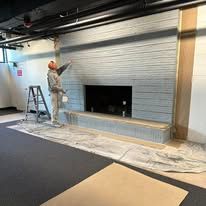 A person stands on a ladder, painting a gray paneled fireplace wall in an unfinished room with protective floor plastic.