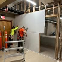 Two workers in high-visibility vests install drywall panels on a wooden frame inside a building under construction.