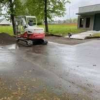 A red and white mini excavator parked on wet asphalt next to a building and a pile of dirt on a rainy day.