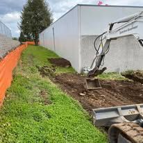 A white excavator is parked on dirt next to a light-colored building, with an orange safety fence along the grassy area.
