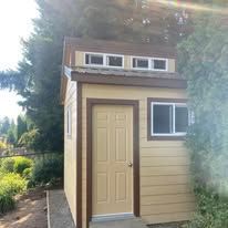 A tan shed with brown trim, a single door, and windows, set outdoors against a backdrop of trees.
