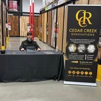 A person sits at a black-covered table in a store, next to a vertical banner for Cedar Creek Renovations.