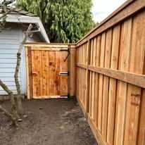 A wooden gate and matching fence corner connected to the side of a house on a dirt surface.