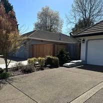 A gray garage with a wood-paneled door, a paved driveway, and shrubs in front under a clear blue sky.