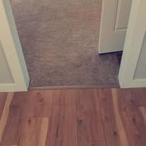 A threshold transition strip connects light-brown wood-look flooring to a doorway leading into a carpeted room.