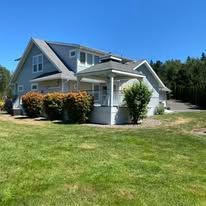 A light blue two-story house with a white gazebo attached to the side, surrounded by a lawn and dense trees.