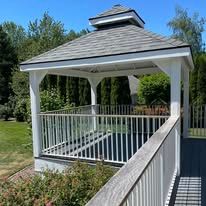A white wooden gazebo with a shingled roof stands on a deck, framed by green trees on a bright, sunny day.
