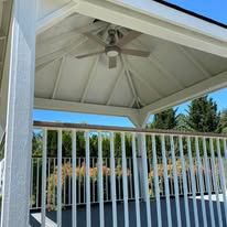White wooden gazebo with a ceiling fan, overlooking a yard with green trees and a white railing in the foreground.