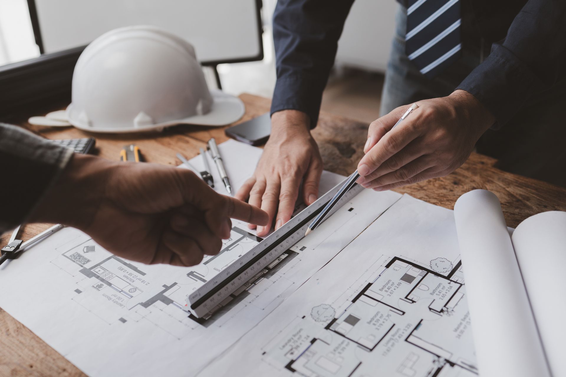 Two people use a drafting scale to review architectural blueprints on a wooden desk with a hard hat nearby.
