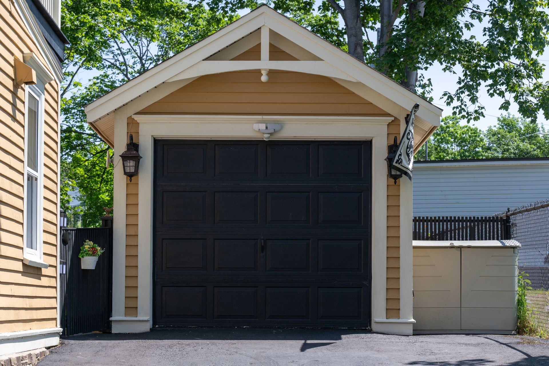 A detached, tan-sided garage with a dark, paneled door and a gabled roof, nestled next to a house on a paved driveway.