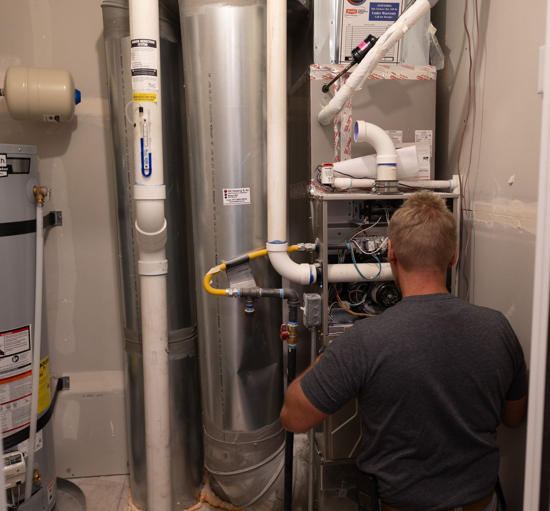Man working on a furnace in a utility room with pipes and water heater; he's wearing a gray shirt and khaki pants.