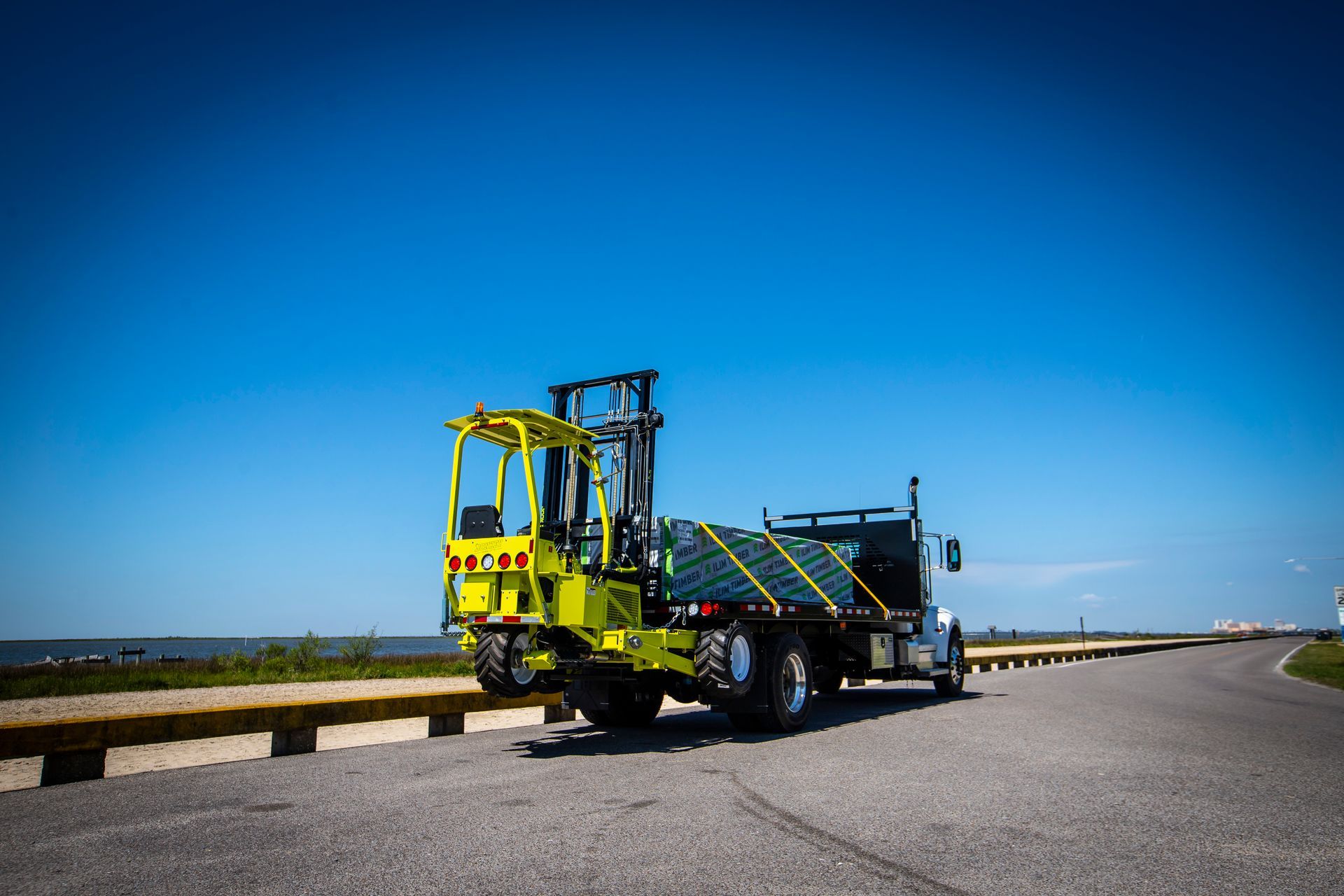 Yellow forklift on a flatbed truck on a road under a blue sky, near a body of water.
