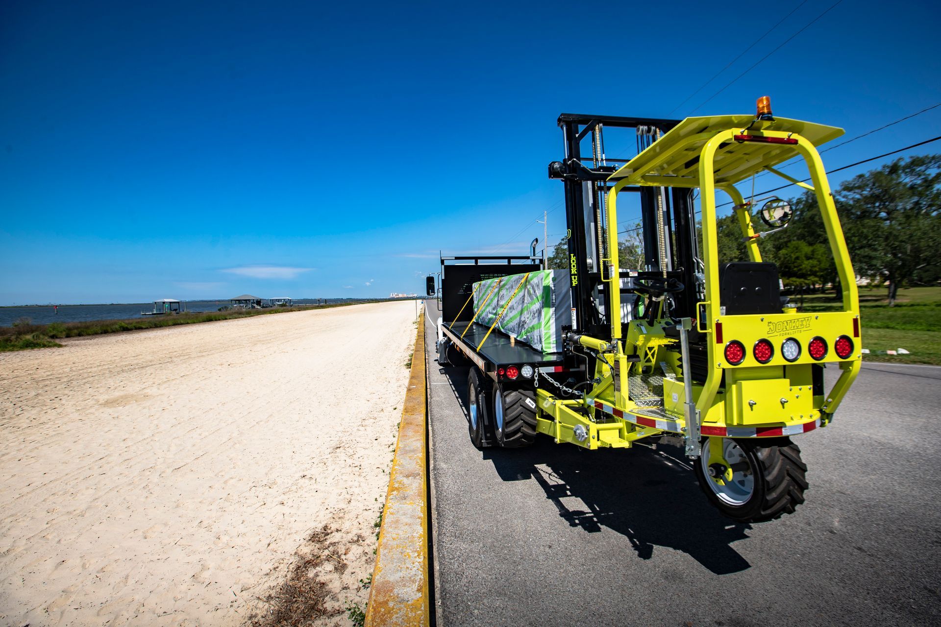 Yellow forklift on a flatbed truck parked next to a sandy beach under a blue sky.