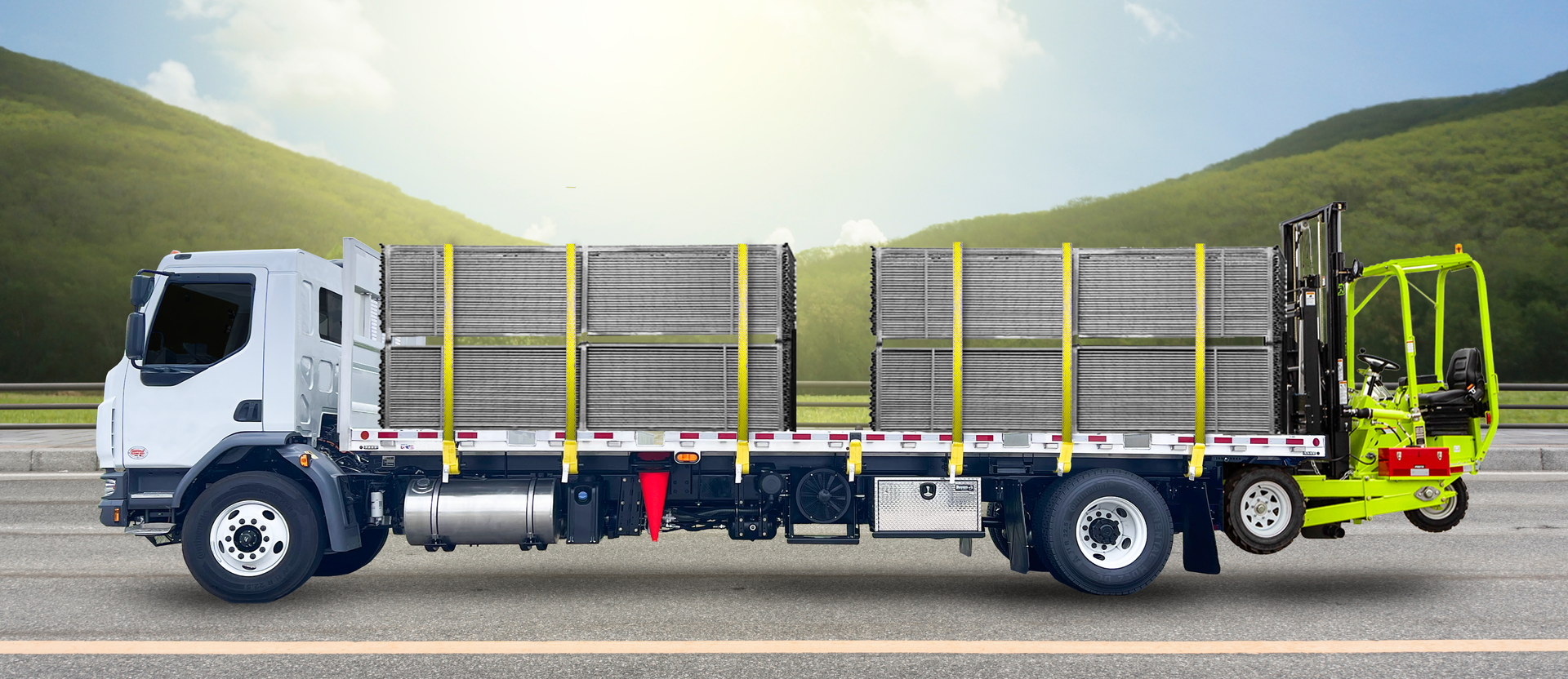 A white truck with cargo strapped down, a green forklift on the back, driving on a road with a green hill.