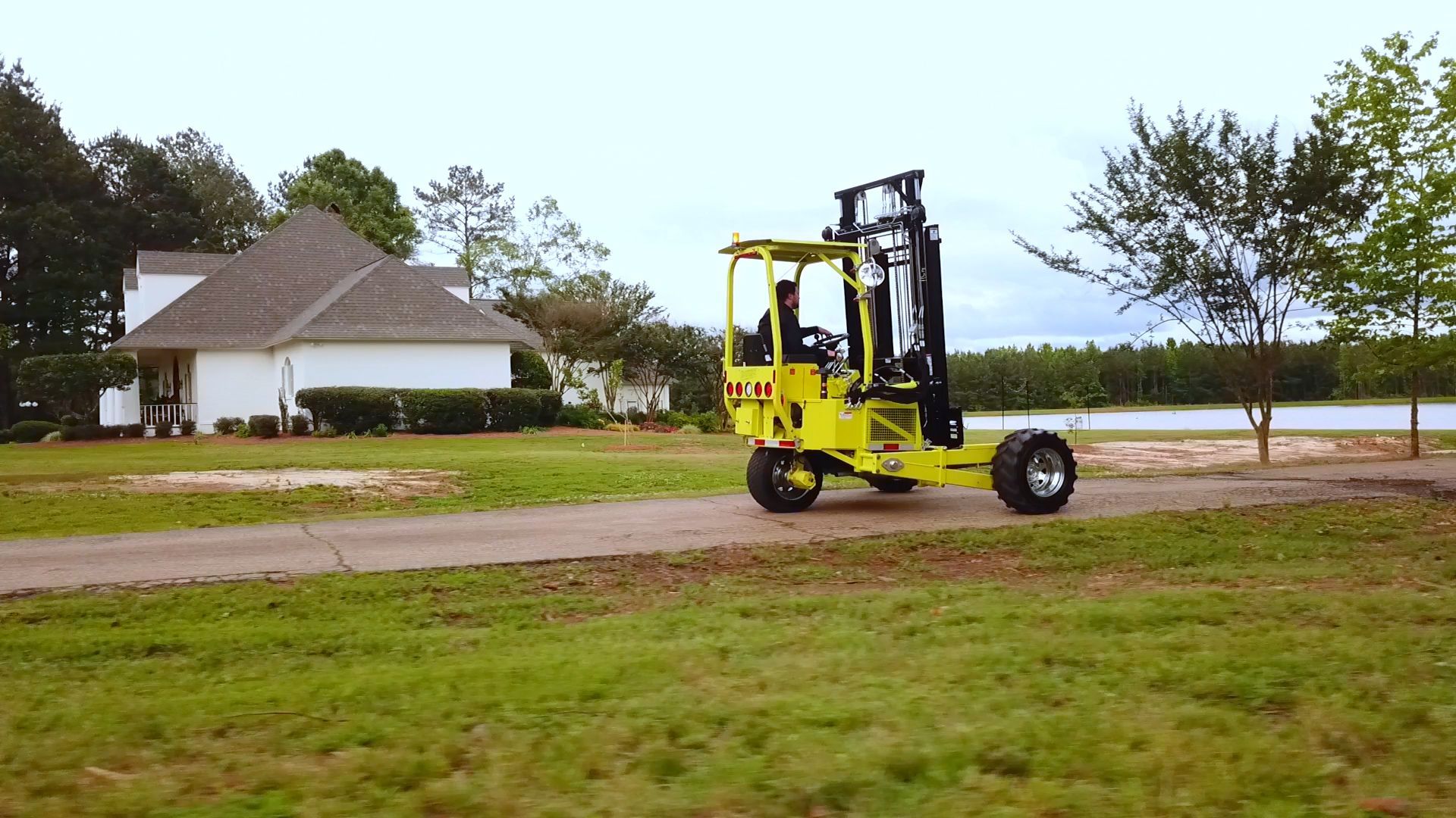 Yellow forklift driving on a dirt road, next to a house and grassy field.