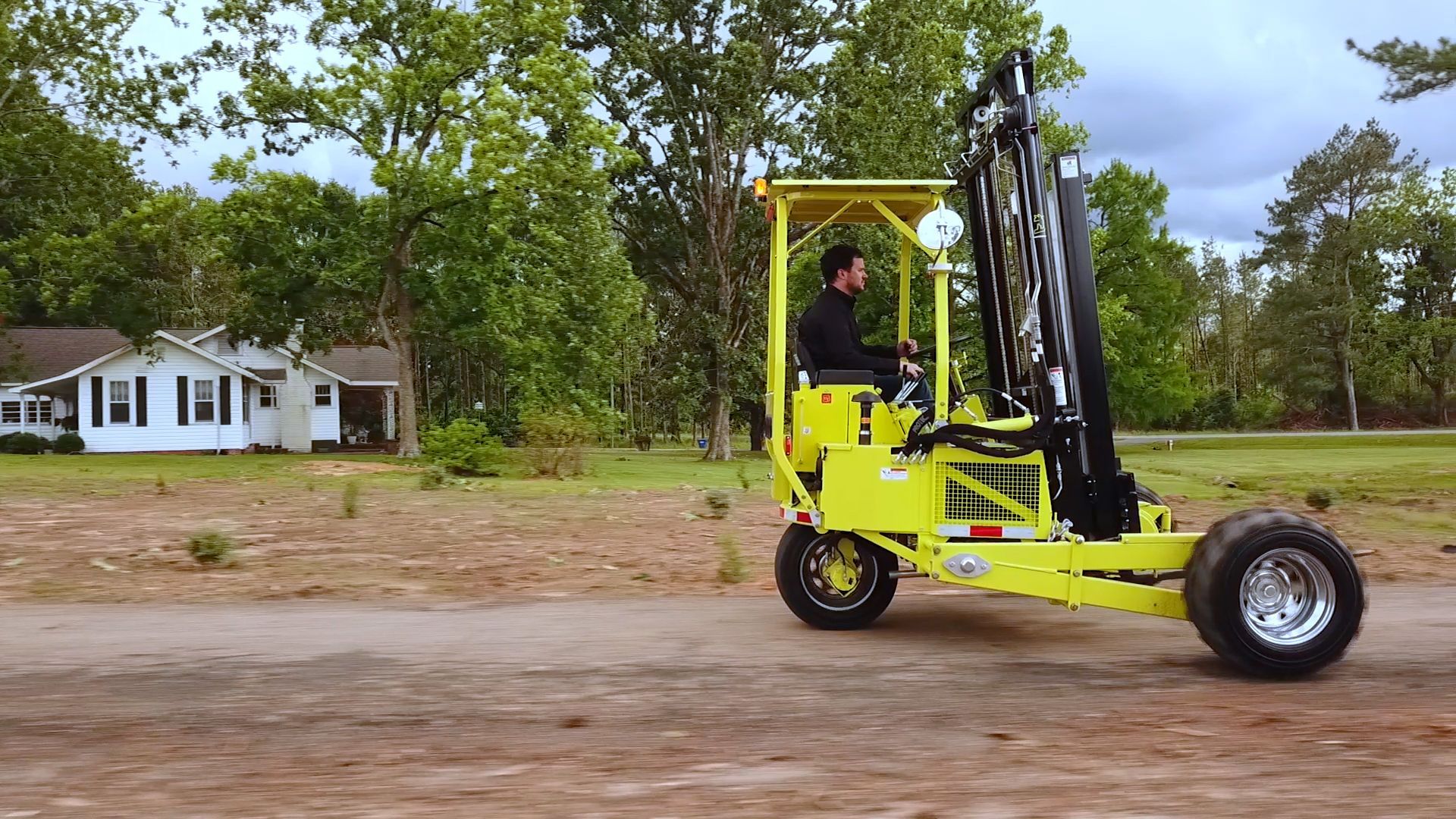 Yellow forklift with driver on dirt road, a house in the background.