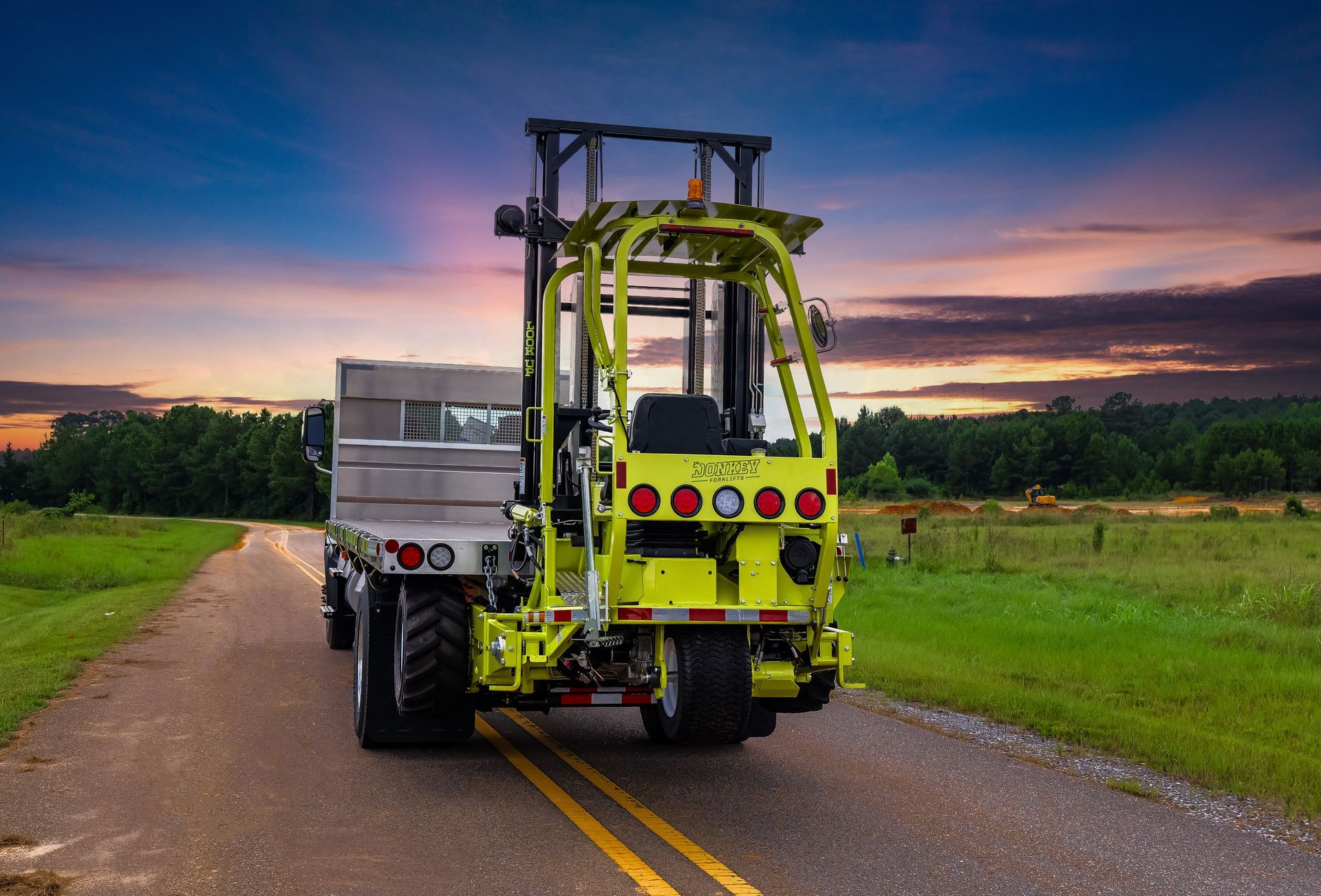 Yellow forklift on a flatbed truck on a rural road at sunset.