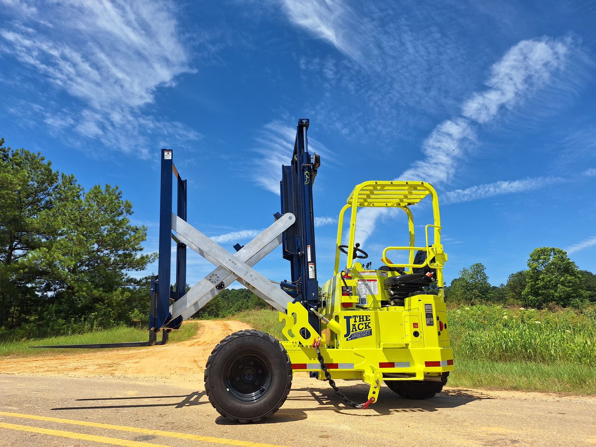 Yellow and blue rough terrain forklift on a gravel road against a blue sky with clouds.