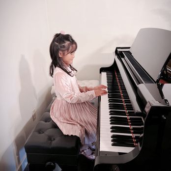 Young girl in pink dress playing a grand piano in a brightly lit room.