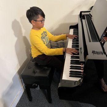 Young person with glasses playing a black piano, sitting on a black bench, in a well-lit room.