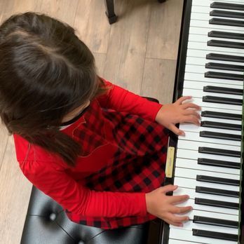 Young girl in red plaid dress playing a piano, seated on a black bench.