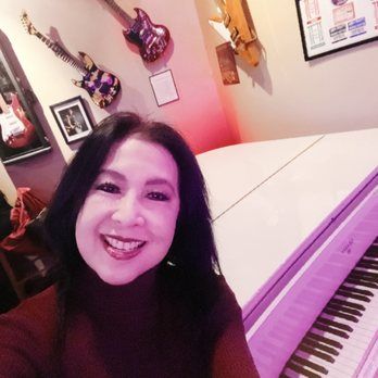 Woman smiling by a white piano, guitars on the wall.  Wearing a red top, in a music-themed space.