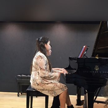 Woman playing a grand piano in a recital setting; she wears a patterned dress.