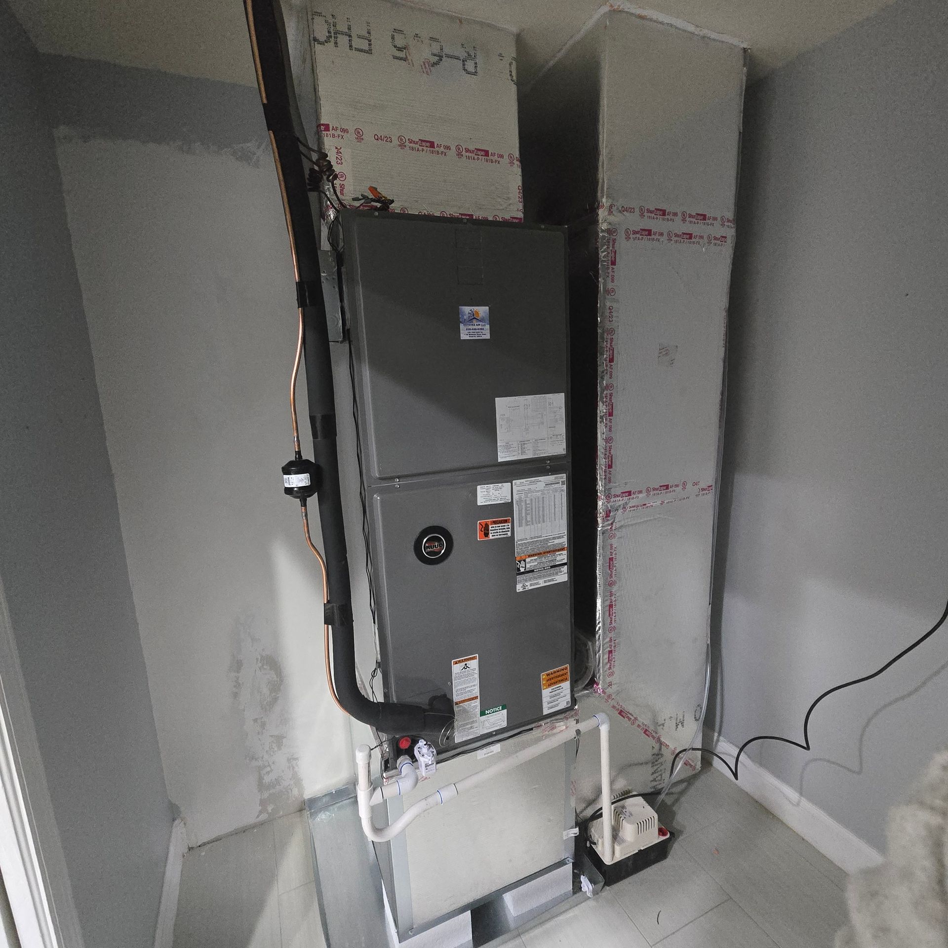 Furnace and ductwork in a utility room. Gray metal unit with pipes and wiring against gray walls.