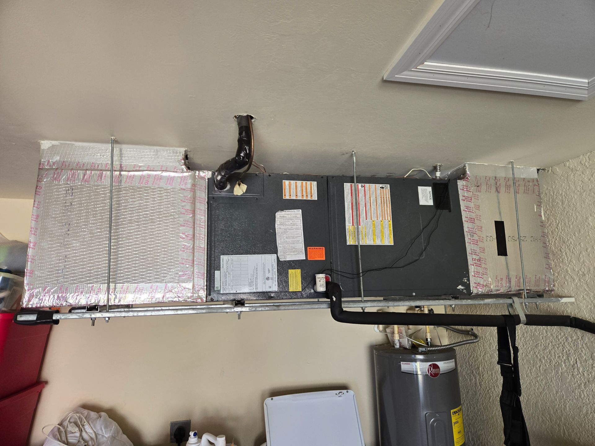 HVAC unit suspended from ceiling in a garage. Ductwork, filter, and water heater are visible.